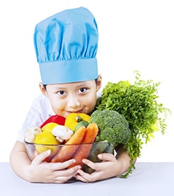 A young boy wearing a chef hat and holding on to a big bowl of vegetables